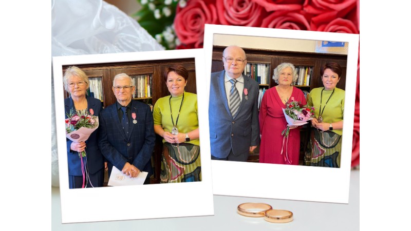 Fotografia przedstawia dwie pary jubilatów odznaczone medalami za długoletnie pożycie małżeńskie wraz z burmistrz miasta Cieszyna, pozujące we wnętrzu z drewnianymi regałami i kwiatami w dłoniach.